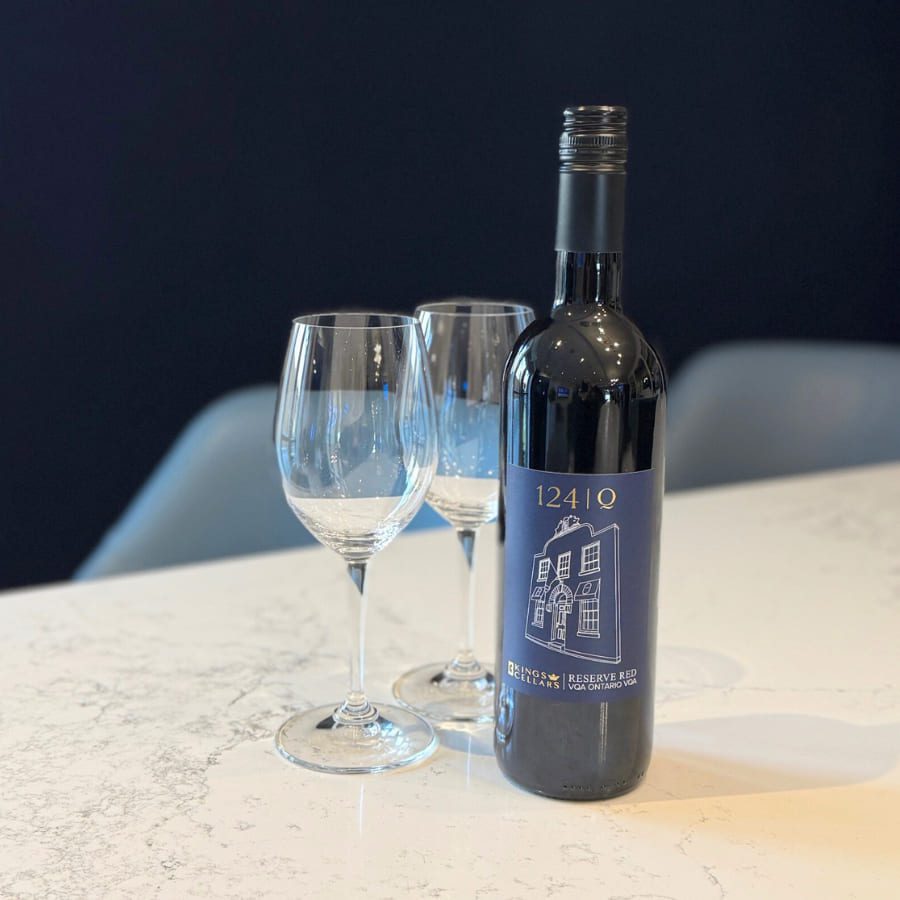 A bottle of red wine labeled 1241 Q stands on a white marble table next to two empty wine glasses. The background is dark, creating a contrast with the bright surface. 124 on Queen Hotel and Spa | Hotel & Spa Niagara-on-the-lake | 1 (855) 988-4552
