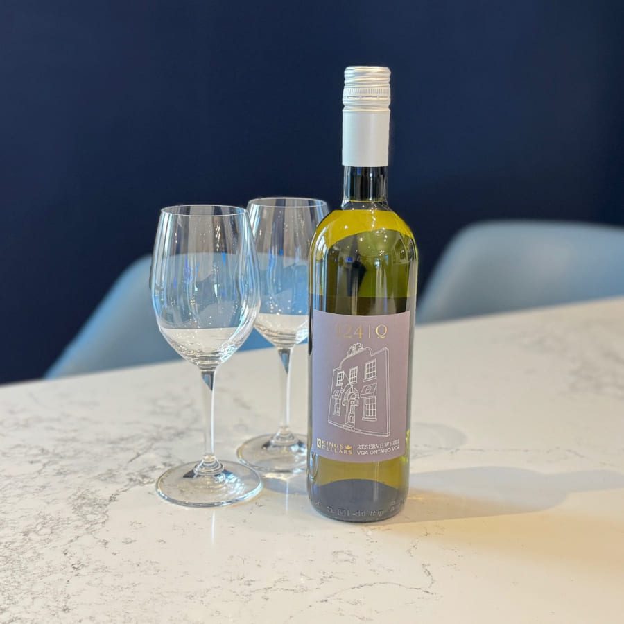A bottle of white wine with a purple label stands on a marble countertop. Two empty wine glasses are beside it. The background is a dark navy blue wall. 124 on Queen Hotel and Spa | Hotel & Spa Niagara-on-the-lake | 1 (855) 988-4552