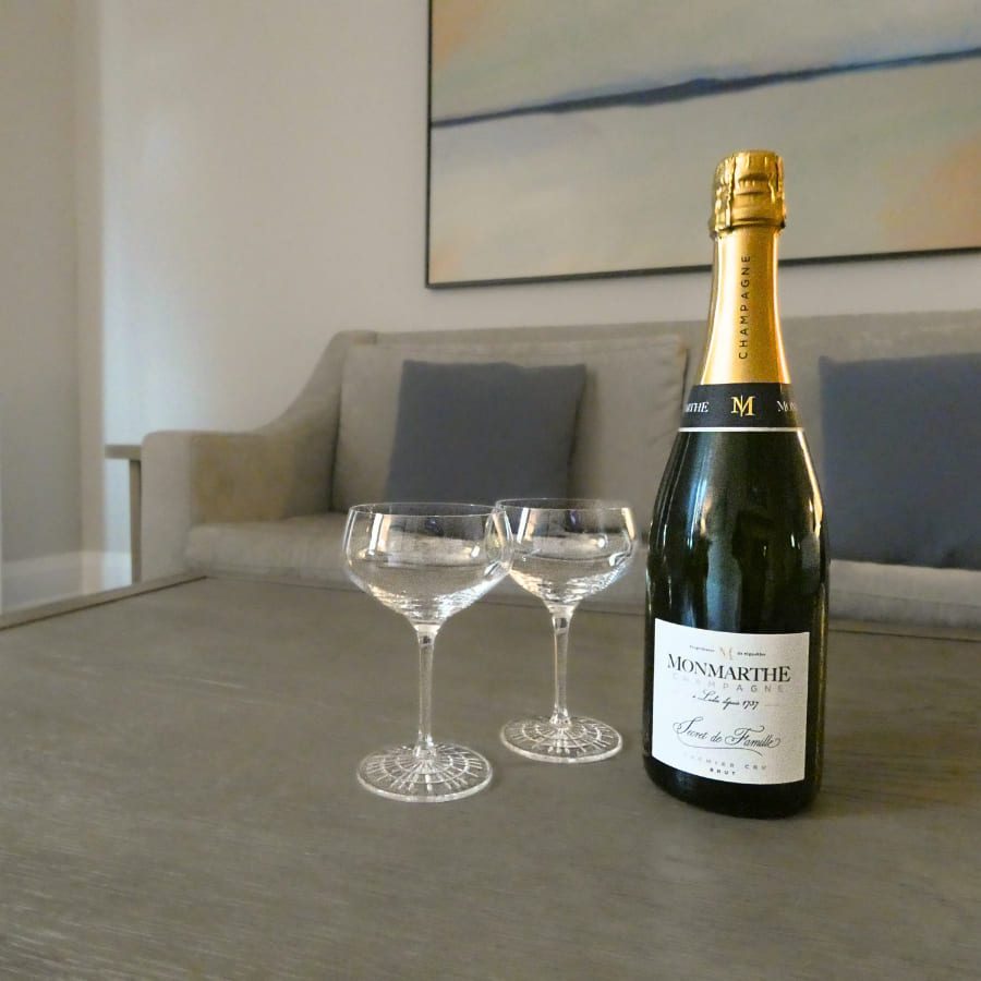 A bottle of champagne and two empty glasses sit on a wooden coffee table. A gray sofa with blue cushions is in the background, along with a large abstract painting on the wall. 124 on Queen Hotel and Spa | Hotel & Spa Niagara-on-the-lake | 1 (855) 988-4552