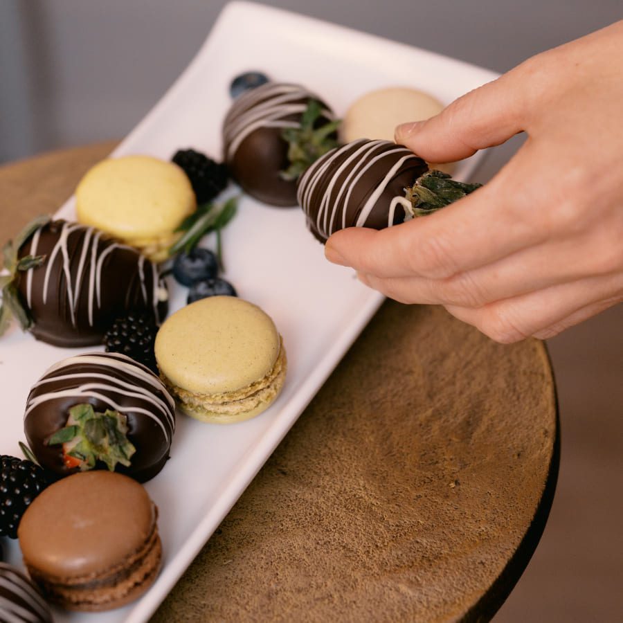 A hand picks up a chocolate-covered strawberry from a white platter containing assorted desserts, including macarons in yellow and brown, and blueberries. The platter is placed on a round wooden table. 124 on Queen Hotel and Spa | Hotel & Spa Niagara-on-the-lake | 1 (855) 988-4552