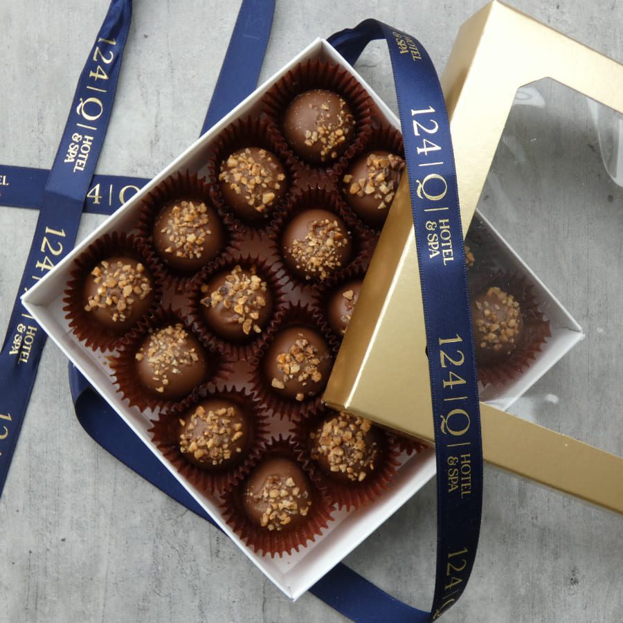 A box of chocolate truffles with nut toppings is open, revealing 16 neatly arranged pieces. The white and gold box is decorated with a blue ribbon printed with 124Q Hotel & Spa. The box rests on a gray surface. 124 on Queen Hotel and Spa | Hotel & Spa Niagara-on-the-lake | 1 (855) 988-4552