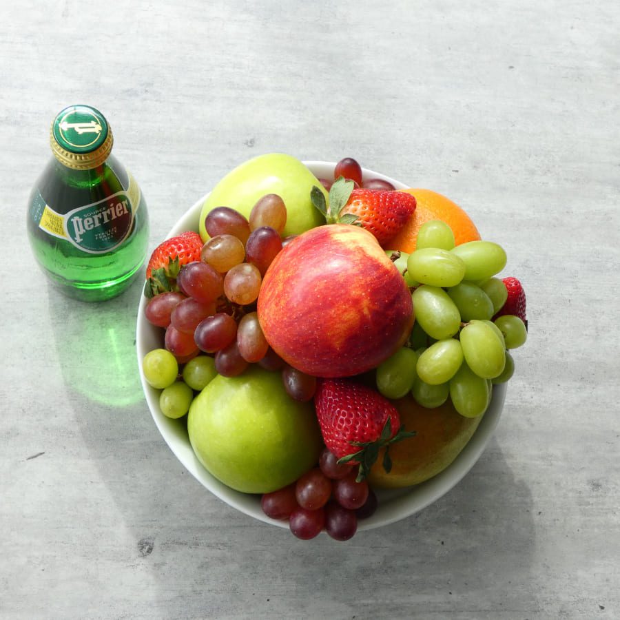 A bowl filled with a variety of fruits, including apples, grapes, strawberries, and an orange, sits on a light gray surface. Beside it is a small bottle of Perrier sparkling water. 124 on Queen Hotel and Spa | Hotel & Spa Niagara-on-the-lake | 1 (855) 988-4552