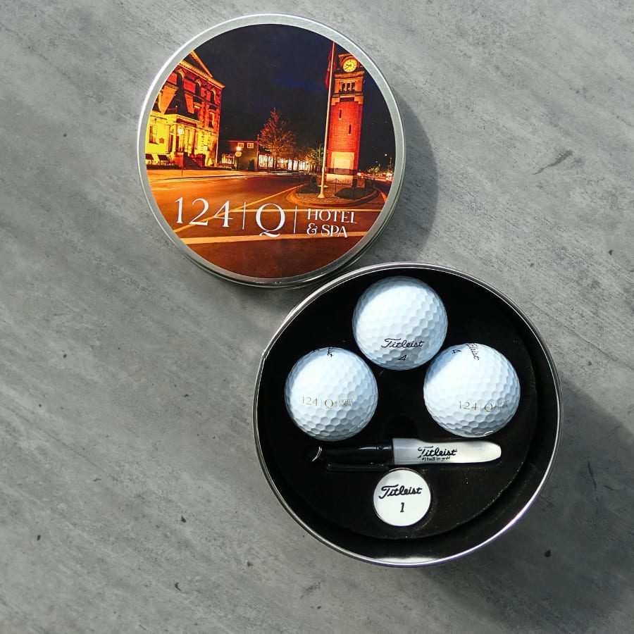 A round tin labeled 124 on Queen Hotel & Spa with a night scene of a building. Inside are three Titleist golf balls and a marker with the same hotel branding, set on a gray surface. 124 on Queen Hotel and Spa | Hotel & Spa Niagara-on-the-lake | 1 (855) 988-4552