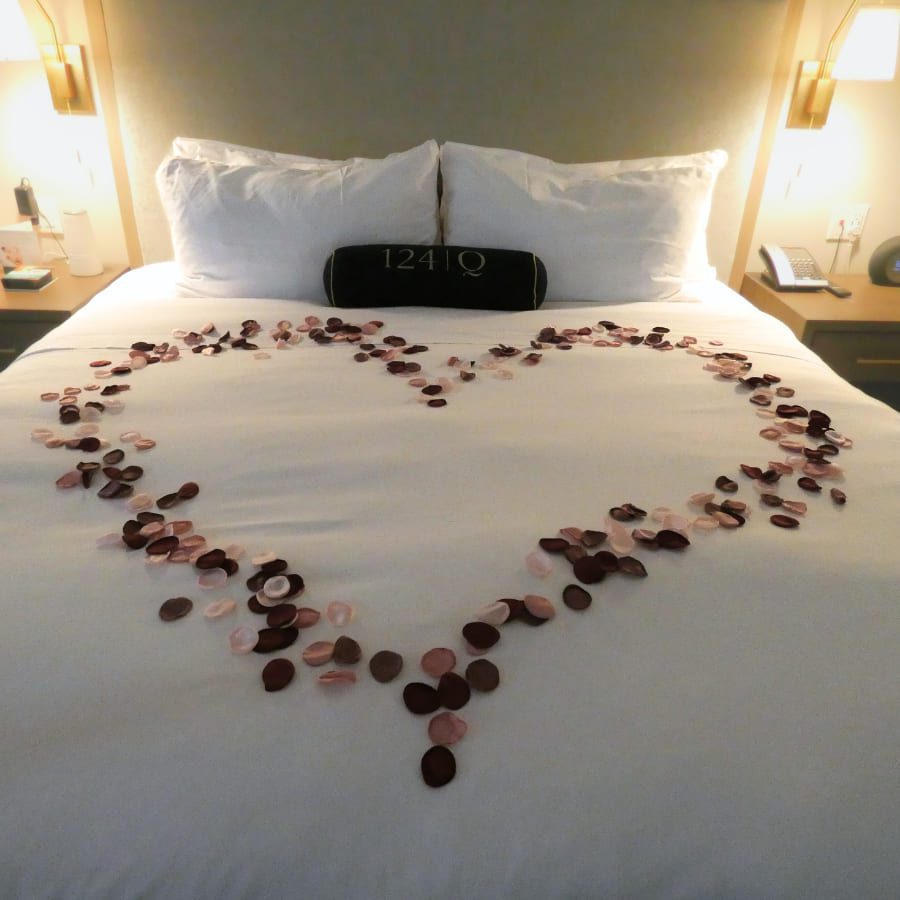A bed is decorated with rose petals arranged in the shape of a heart. The bedding is white, and there is a dark pillow with a number on it resting against the headboard. Soft lighting creates a cozy ambiance. 124 on Queen Hotel and Spa | Hotel & Spa Niagara-on-the-lake | 1 (855) 988-4552