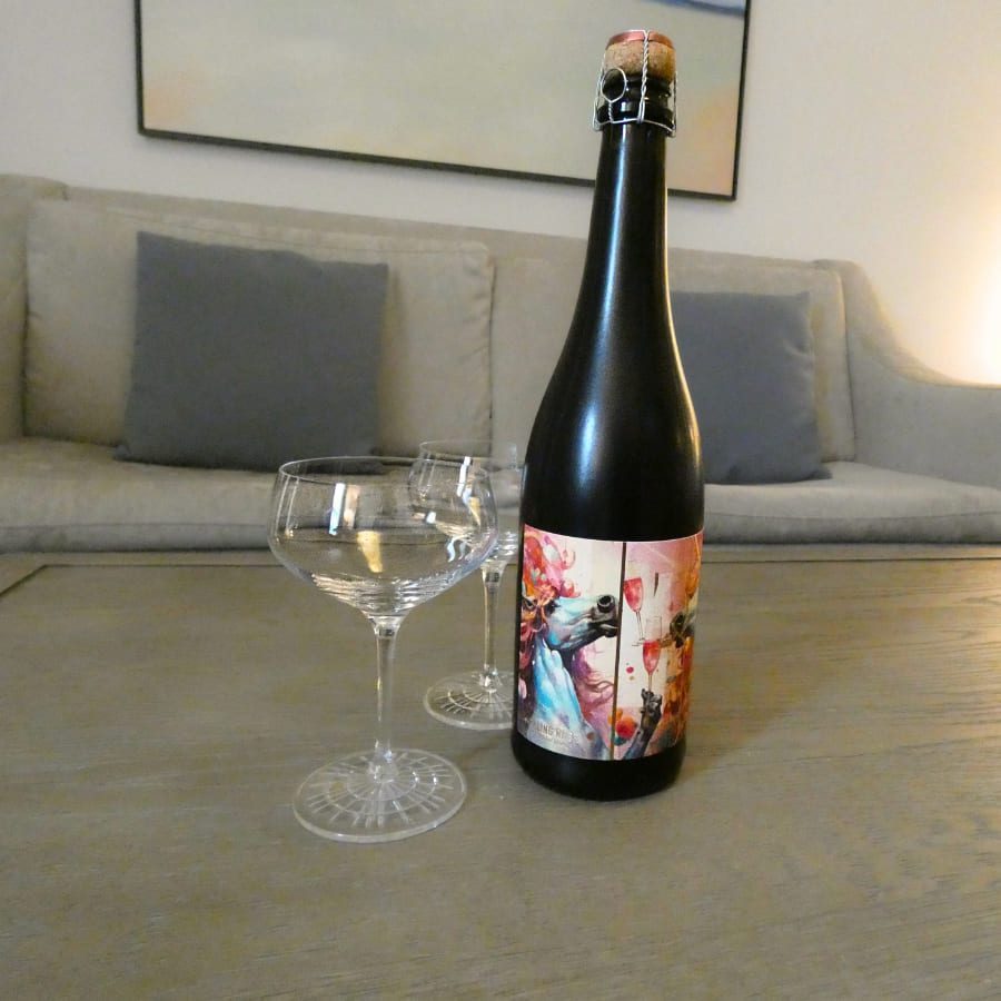 A black bottle with an artistic label stands on a wooden table, accompanied by two clear, elegant glasses. Behind them is a light-colored couch with gray cushions, under a large painting. The scene is warmly lit, creating a cozy ambiance. 124 on Queen Hotel and Spa | Hotel & Spa Niagara-on-the-lake | 1 (855) 988-4552