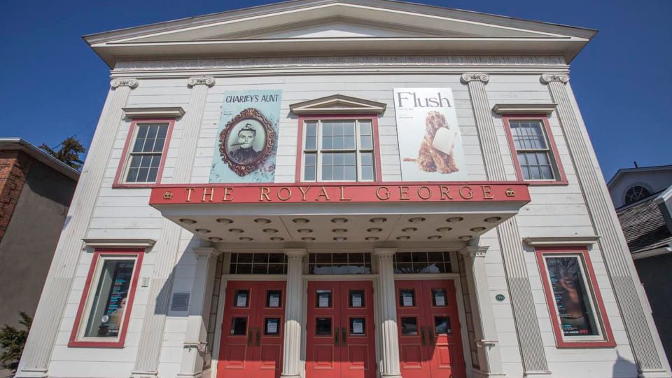 Front view of The Royal George Theatre, a white building with a red marquee. Posters for Charleys Aunt and Flush are displayed above the entrance, which features several red doors. The sky is clear and blue.