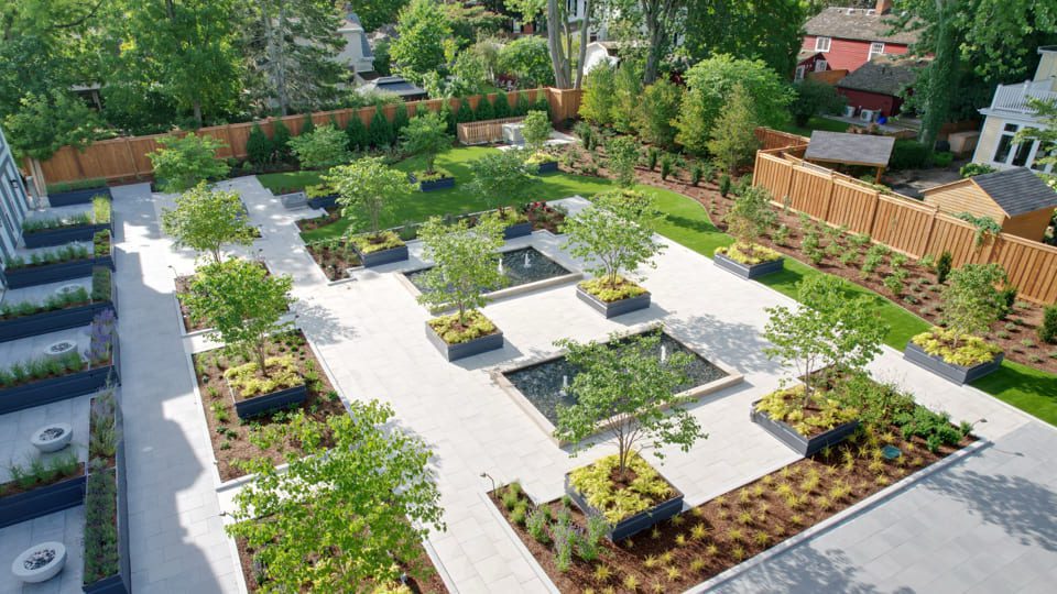 A landscaped garden with geometric paved paths and planters, featuring small trees and two rectangular reflecting pools. The area is surrounded by wooden fencing, with lush greenery and residential buildings in the background.