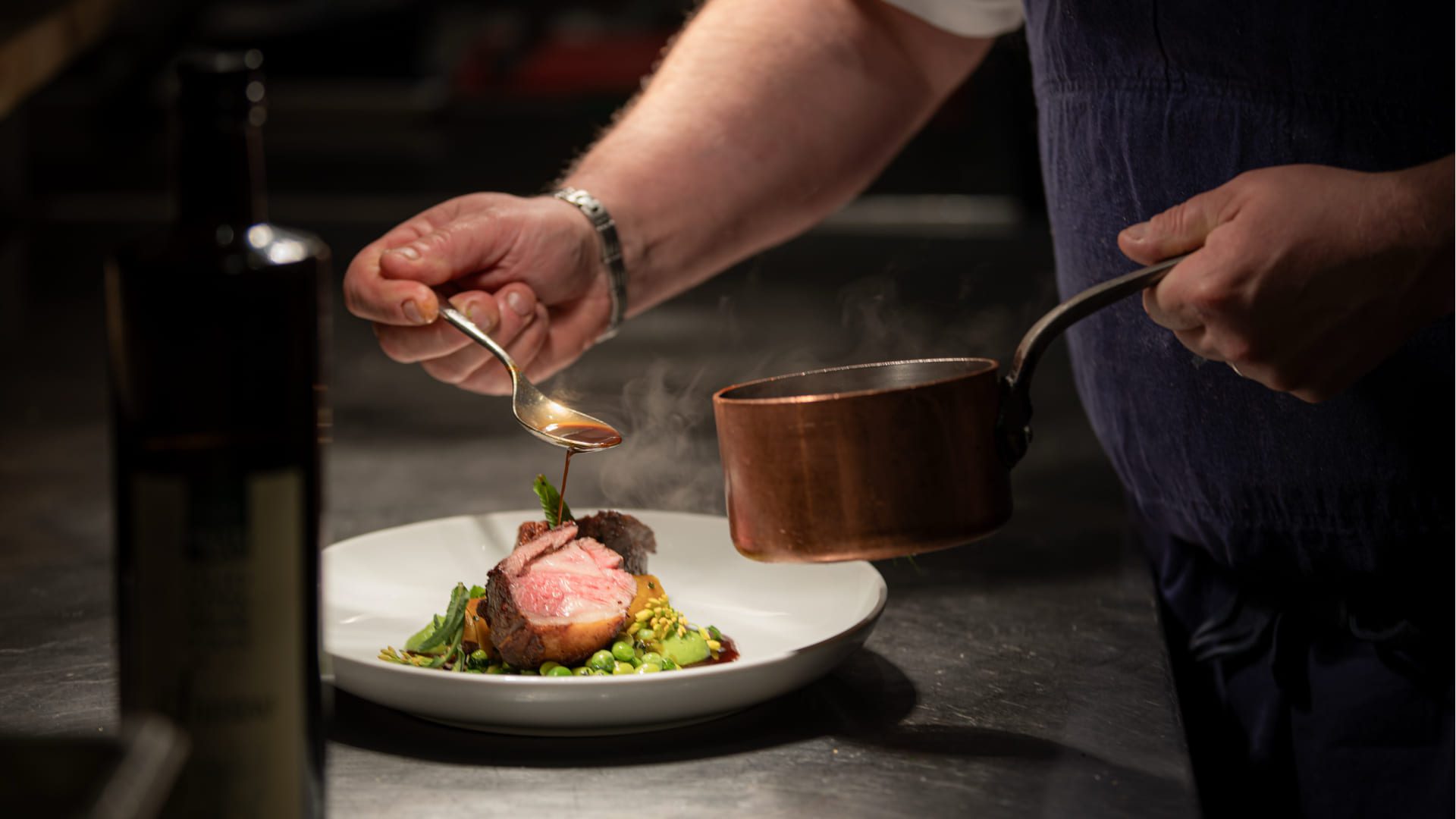 A chef pours sauce from a copper pot onto a plated dish of sliced meat and vegetables, with steam rising from the food in a dimly lit kitchen. 124 on Queen Hotel and Spa | Hotel & Spa Niagara-on-the-lake | 1 (855) 988-4552