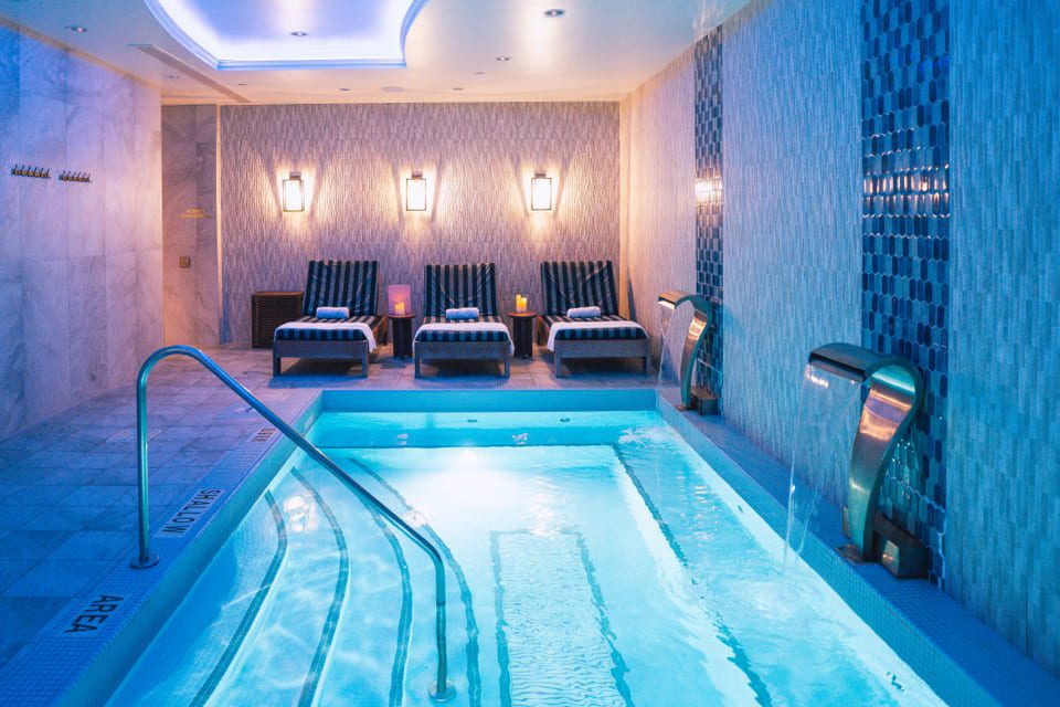 A luxurious indoor spa room with a glowing blue pool, water features, and four cushioned lounge chairs lined up along the back wall, illuminated by warm wall lights. 124 on Queen Hotel and Spa | Hotel & Spa Niagara-on-the-lake | 1 (855) 988-4552
