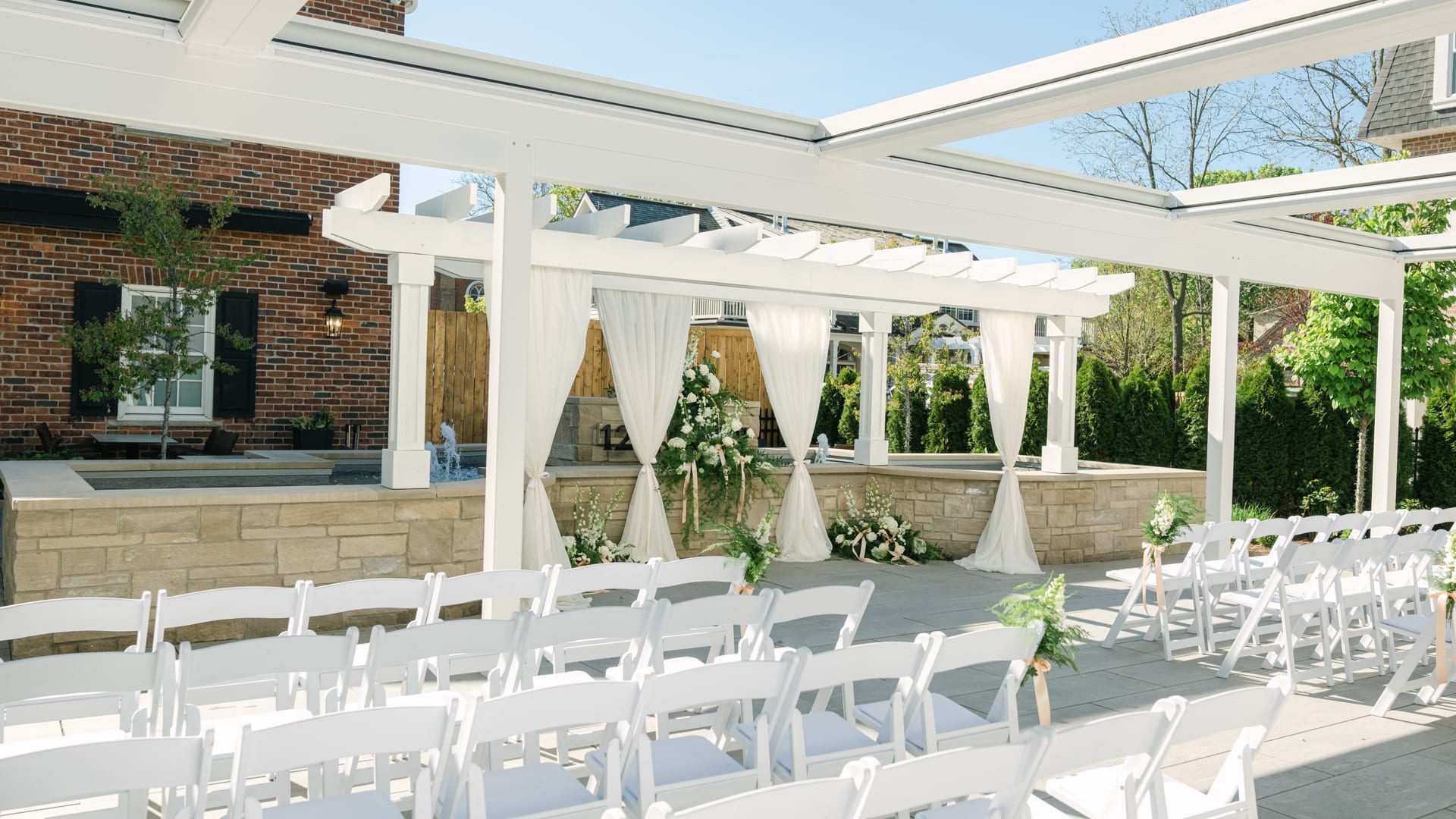 An outdoor wedding ceremony setup with white folding chairs facing a pergola decorated with white drapes and floral arrangements, set on a sunny patio beside a brick building. 124 on Queen Hotel and Spa | Hotel & Spa Niagara-on-the-lake | 1 (855) 988-4552