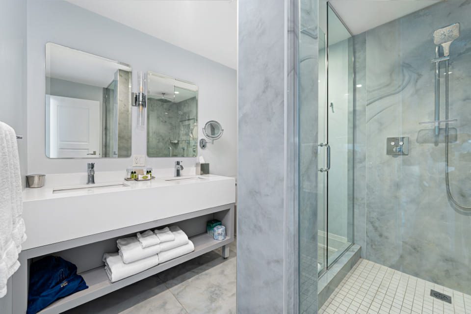Modern bathroom with a double sink vanity, large mirrors, neatly folded towels on open shelves, and a glass-enclosed shower with a rainfall showerhead and gray marble walls. 124 on Queen Hotel and Spa | Hotel & Spa Niagara-on-the-lake | 1 (855) 988-4552