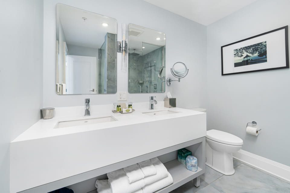 Modern bathroom with a double sink vanity, two mirrors, neatly arranged toiletries, a wall-mounted toilet, a glass-enclosed shower, towels on a shelf, and framed art on a light blue wall. 124 on Queen Hotel and Spa | Hotel & Spa Niagara-on-the-lake | 1 (855) 988-4552