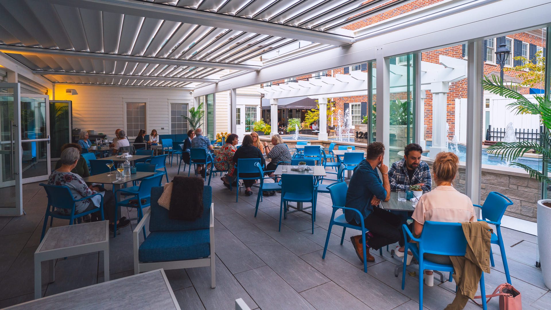 People are dining and conversing at tables in a modern, covered outdoor patio with blue chairs, glass walls, and plenty of natural light, enjoying late summer food and wine. The atmosphere is casual and relaxed in Niagara-on-the-Lake. 124 on Queen Hotel and Spa | Hotel & Spa Niagara-on-the-lake | 1 (855) 988-4552
