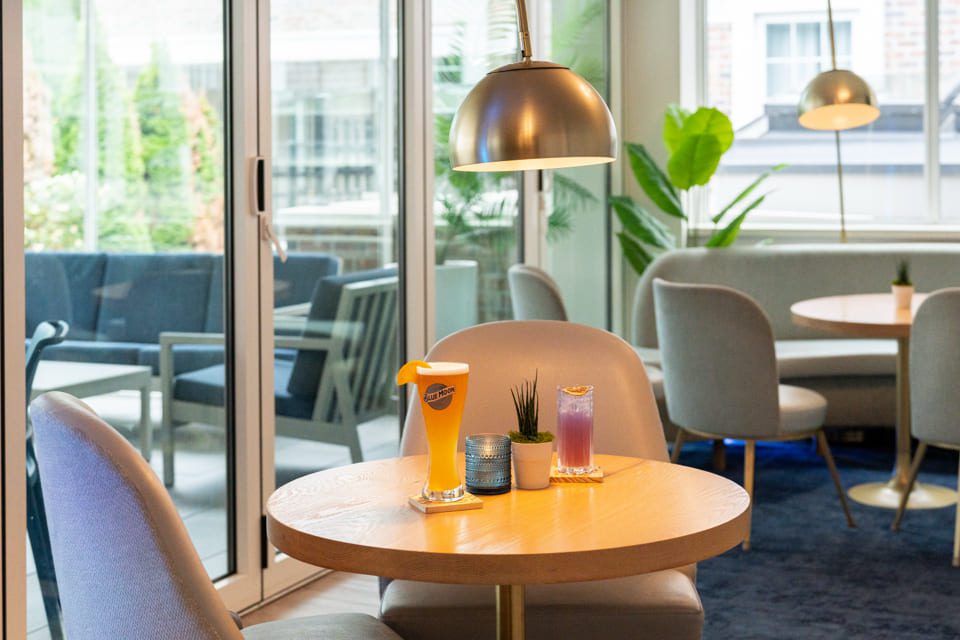 A modern café with light gray chairs and a round wooden table set with two colorful drinks and small plants, next to large windows overlooking an outdoor seating area with greenery. 124 on Queen Hotel and Spa | Hotel & Spa Niagara-on-the-lake | 1 (855) 988-4552