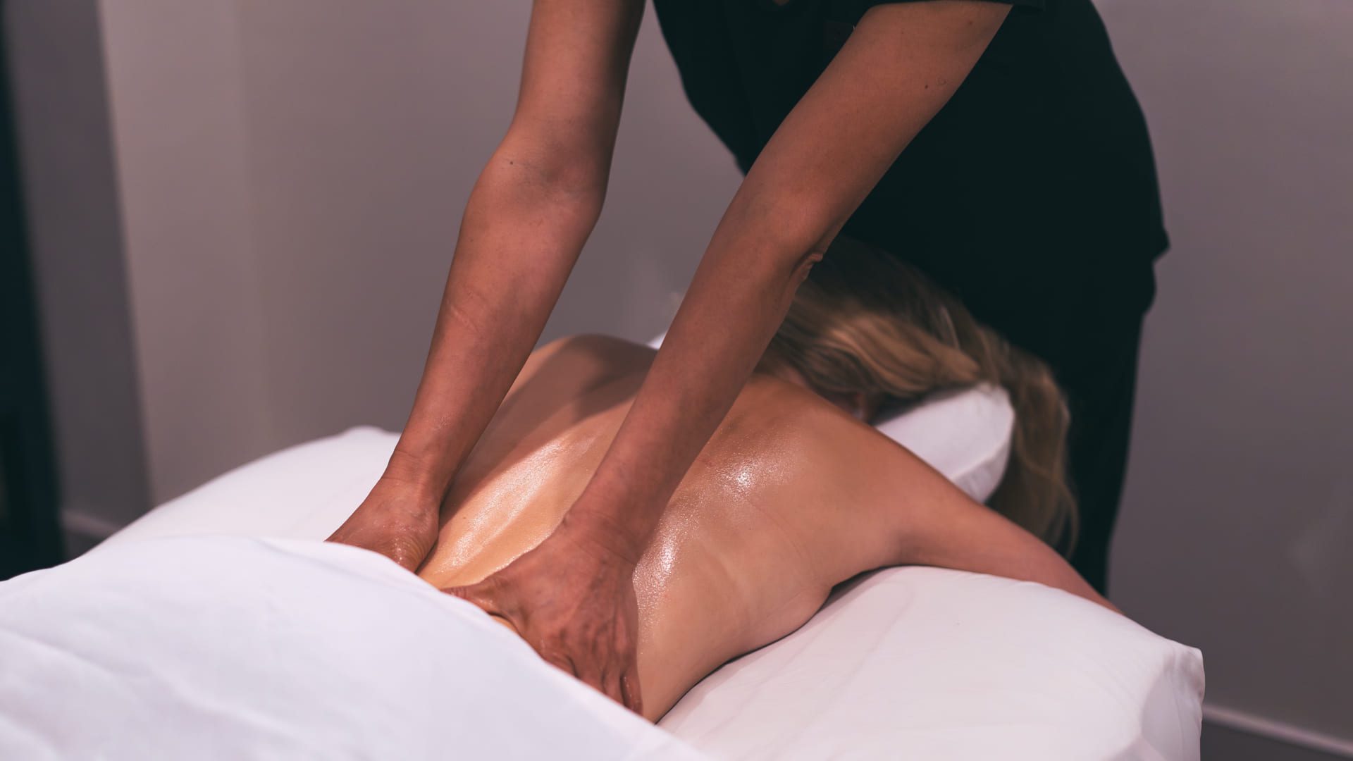A person receives a back massage while lying face down on a massage table, covered with a white sheet. The masseuse uses both hands to apply pressure to the client’s back. 124 on Queen Hotel and Spa | Hotel & Spa Niagara-on-the-lake | 1 (855) 988-4552