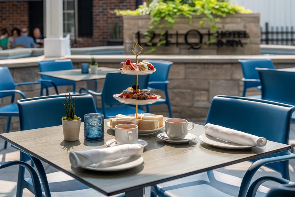 A set table for two outdoors with blue chairs, white napkins, teacups, and a tiered tray of pastries and sandwiches. The background shows a hotel sign and greenery. 124 on Queen Hotel and Spa | Hotel & Spa Niagara-on-the-lake | 1 (855) 988-4552