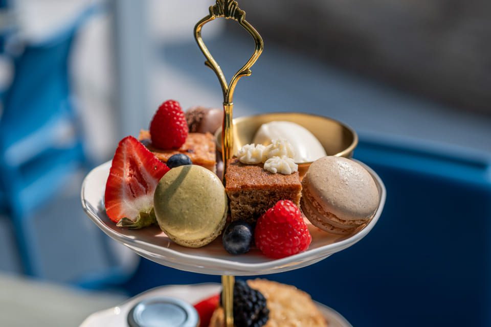 A two-tiered dessert stand holds assorted pastries, including macarons, a slice of cake, fresh berries, and a bowl of cream, with a blurred blue chair and table in the background. 124 on Queen Hotel and Spa | Hotel & Spa Niagara-on-the-lake | 1 (855) 988-4552