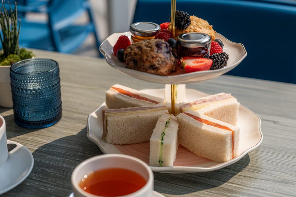 A tiered serving tray with assorted finger sandwiches on the bottom and scones, berries, and jam jars on top, beside a cup of tea and a blue glass on a wooden table. 124 on Queen Hotel and Spa | Hotel & Spa Niagara-on-the-lake | 1 (855) 988-4552