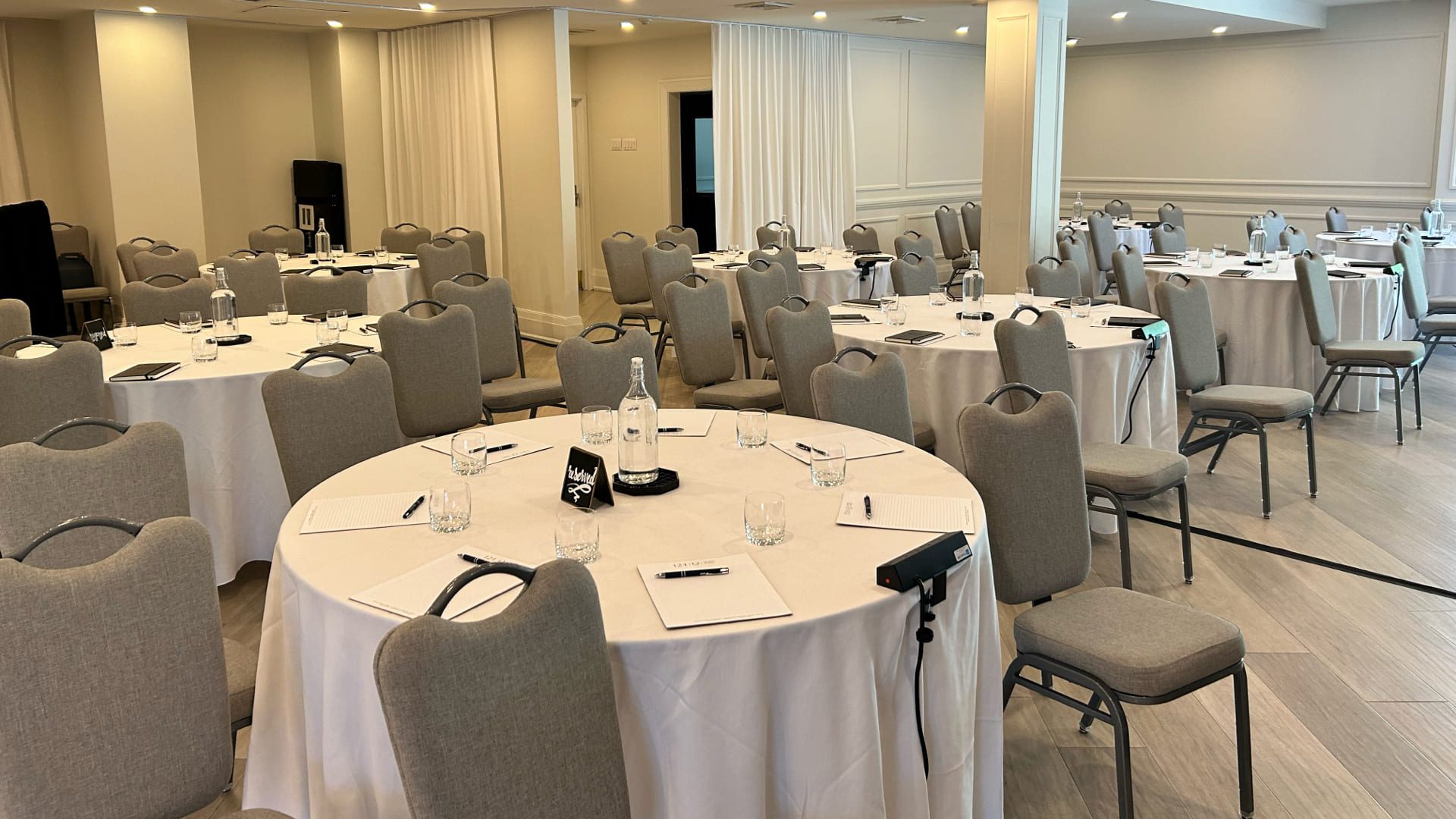 A conference room with round tables covered in white tablecloths, each set with notepads, pens, water bottles, and glasses. Gray chairs surround the tables and the room is well-lit with neutral decor. 124 on Queen Hotel and Spa | Hotel & Spa Niagara-on-the-lake | 1 (855) 988-4552