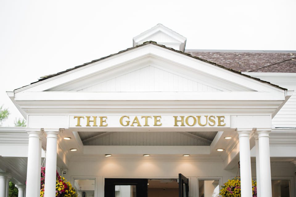 A white building with columns features a gold sign that reads THE GATE HOUSE above the entrance. Hanging flower baskets decorate each side of the entryway. 124 on Queen Hotel and Spa | Hotel & Spa Niagara-on-the-lake | 1 (855) 988-4552