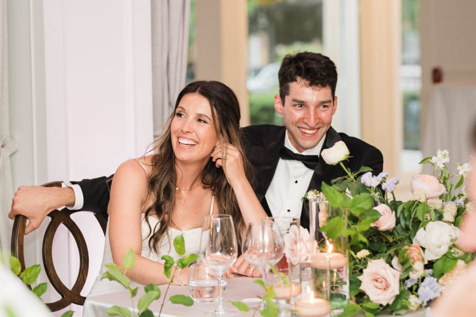 A bride and groom sit smiling together at a table decorated with flowers and candles, dressed in a white wedding gown and black tuxedo, enjoying a joyful moment during their reception. 124 on Queen Hotel and Spa | Hotel & Spa Niagara-on-the-lake | 1 (855) 988-4552