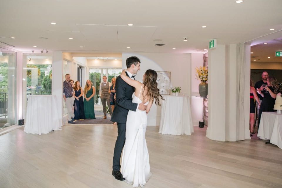 A bride and groom share their first dance in a bright, modern reception hall while guests watch and smile in the background. The room has large windows and light wooden floors. 124 on Queen Hotel and Spa | Hotel & Spa Niagara-on-the-lake | 1 (855) 988-4552