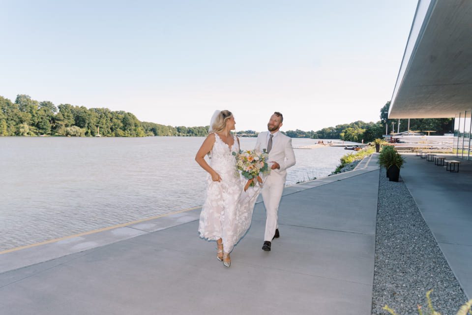 A bride in a white dress holds a bouquet and smiles at a groom in a light suit as they walk together along a waterfront, with trees and blue sky in the background. 124 on Queen Hotel and Spa | Hotel & Spa Niagara-on-the-lake | 1 (855) 988-4552