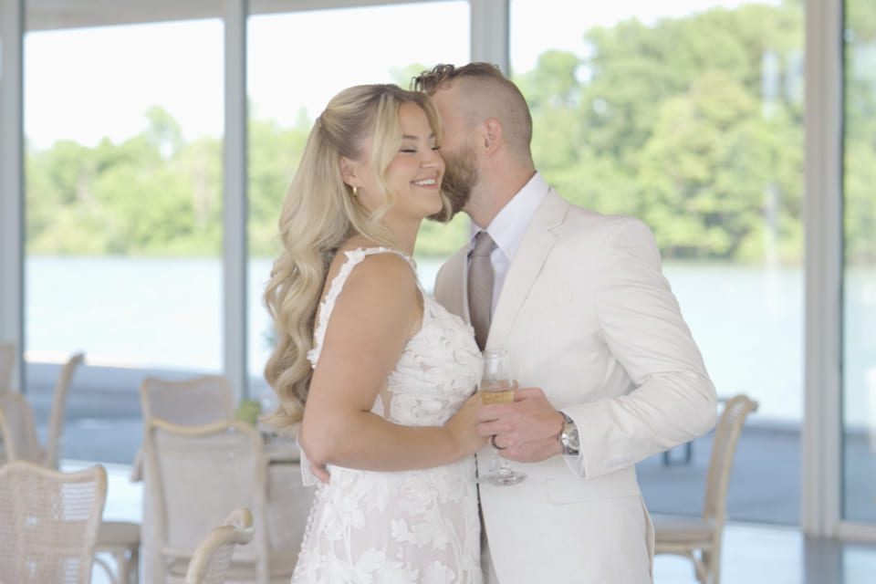A smiling bride in a white dress holds a glass of champagne as a groom in a light suit kisses her cheek. They stand indoors by large windows with a scenic view of trees and water in the background. 124 on Queen Hotel and Spa | Hotel & Spa Niagara-on-the-lake | 1 (855) 988-4552