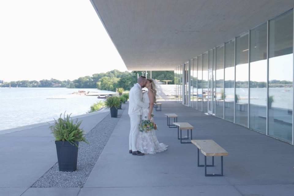 A bride and groom stand close together, facing each other and holding hands, outside a modern building by the water. They are dressed in wedding attire, with glass walls and benches along the walkway. 124 on Queen Hotel and Spa | Hotel & Spa Niagara-on-the-lake | 1 (855) 988-4552