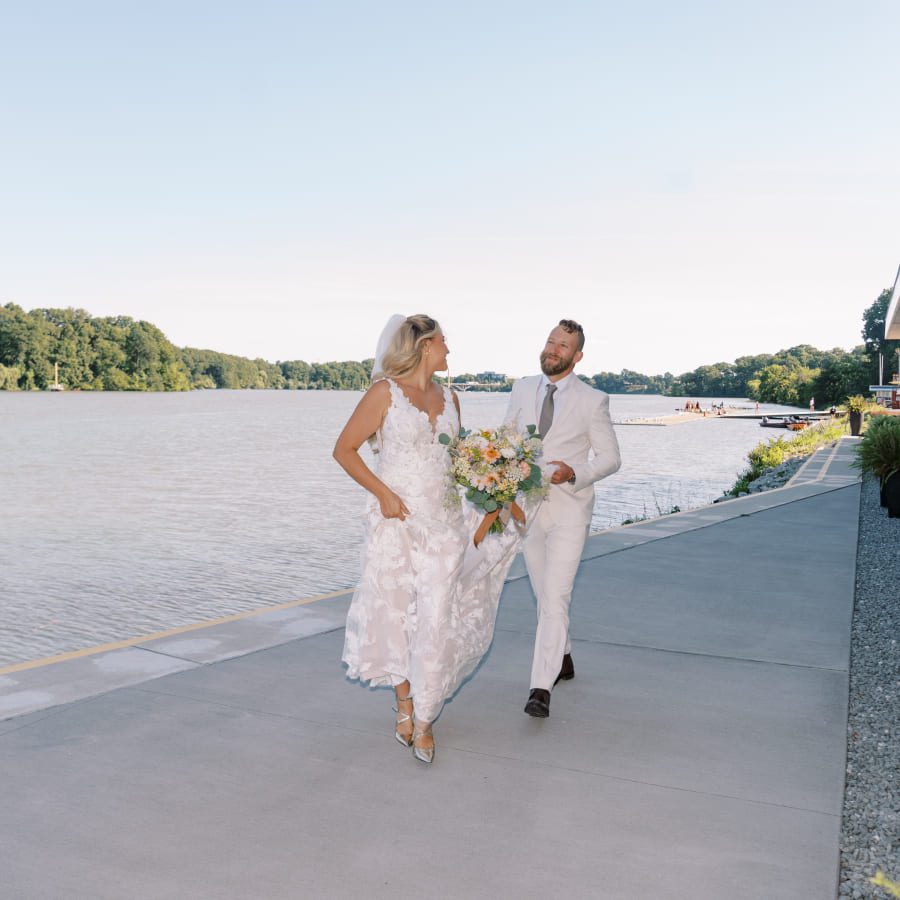 A bride in a white dress holds a bouquet and smiles at a groom in a light suit as they walk together along a waterfront, with trees and blue sky in the background. 124 on Queen Hotel and Spa | Hotel & Spa Niagara-on-the-lake | 1 (855) 988-4552