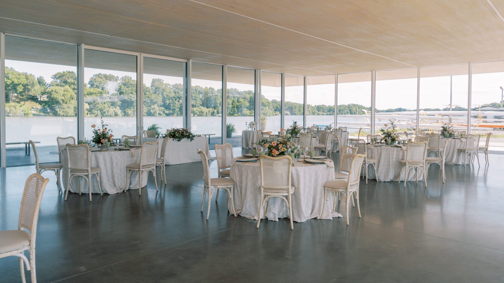 An elegant event space with round tables covered in white linens, floral centerpieces, and white chairs. Large windows provide views of trees and a river outside, filling the room with natural light. 124 on Queen Hotel and Spa | Hotel & Spa Niagara-on-the-lake | 1 (855) 988-4552