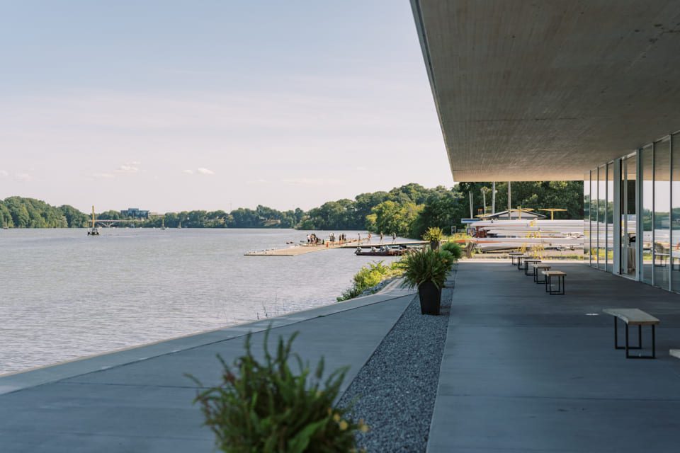 A modern building with large windows and benches overlooks a calm river with docks and people in the distance, surrounded by trees under a clear sky. 124 on Queen Hotel and Spa | Hotel & Spa Niagara-on-the-lake | 1 (855) 988-4552