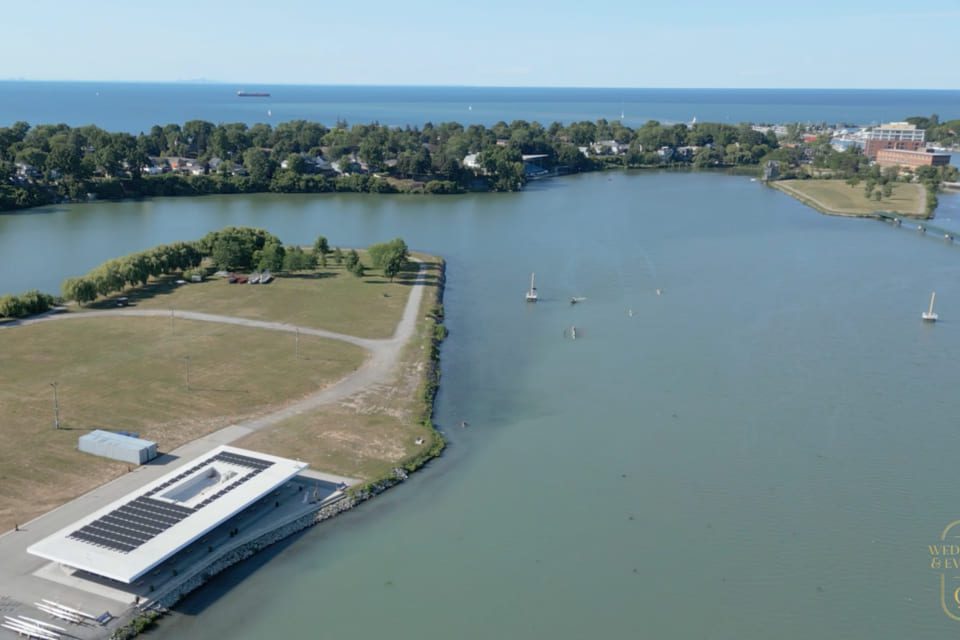 Aerial view of a river meeting a large lake, with sailboats on the water, a modern white building with solar panels on the roof, green trees, open fields, and distant buildings under a clear blue sky. 124 on Queen Hotel and Spa | Hotel & Spa Niagara-on-the-lake | 1 (855) 988-4552