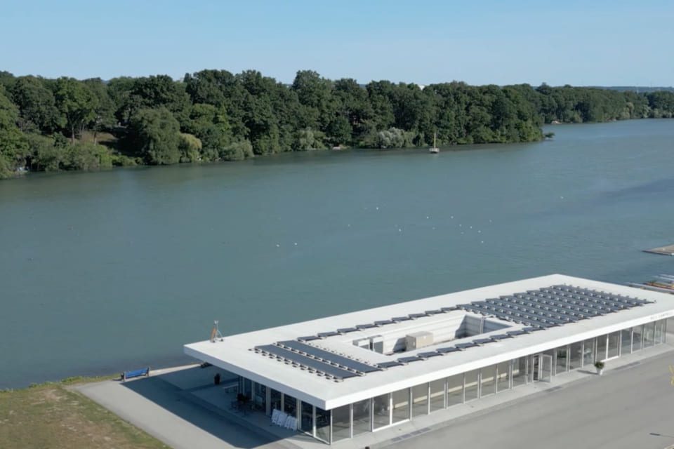 A modern white building with a rooftop solar panel array sits beside a calm river, with dense green trees lining the opposite shore under a clear blue sky. 124 on Queen Hotel and Spa | Hotel & Spa Niagara-on-the-lake | 1 (855) 988-4552