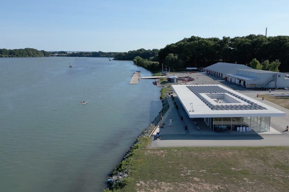 A modern white building stands near the edge of a calm river, surrounded by trees and additional structures. A small boat is visible on the water under a clear blue sky. 124 on Queen Hotel and Spa | Hotel & Spa Niagara-on-the-lake | 1 (855) 988-4552