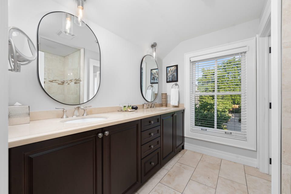 Modern bathroom with a long beige countertop, double sinks, dark wood cabinets, two round mirrors, and a large window with white blinds letting in natural light. Light-colored tile floor and bright, clean decor. 124 on Queen Hotel and Spa | Hotel & Spa Niagara-on-the-lake | 1 (855) 988-4552