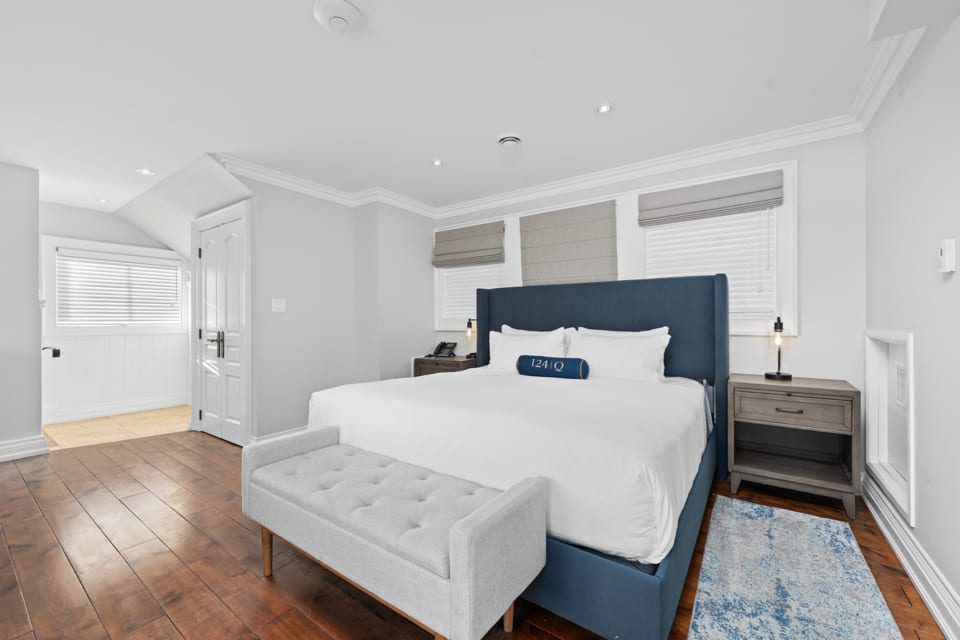 A modern bedroom with a large blue upholstered bed, white bedding, gray bench, two nightstands with lamps, hardwood floors, and three windows with gray shades. The room is bright with white walls and ample natural light. 124 on Queen Hotel and Spa | Hotel & Spa Niagara-on-the-lake | 1 (855) 988-4552