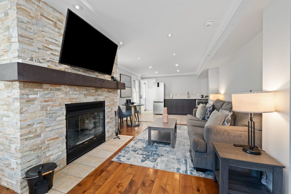 Modern living room with a stone fireplace, wall-mounted TV, gray sofa, blue patterned rug, wooden floors, and an open kitchen and dining area in the background. 124 on Queen Hotel and Spa | Hotel & Spa Niagara-on-the-lake | 1 (855) 988-4552