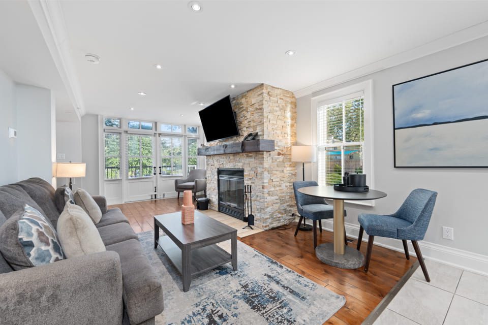 Bright living room with large windows, a gray sofa, modern rug, coffee table, stone fireplace with a mounted TV, and a small round dining table with two blue chairs near a window and abstract wall art. 124 on Queen Hotel and Spa | Hotel & Spa Niagara-on-the-lake | 1 (855) 988-4552