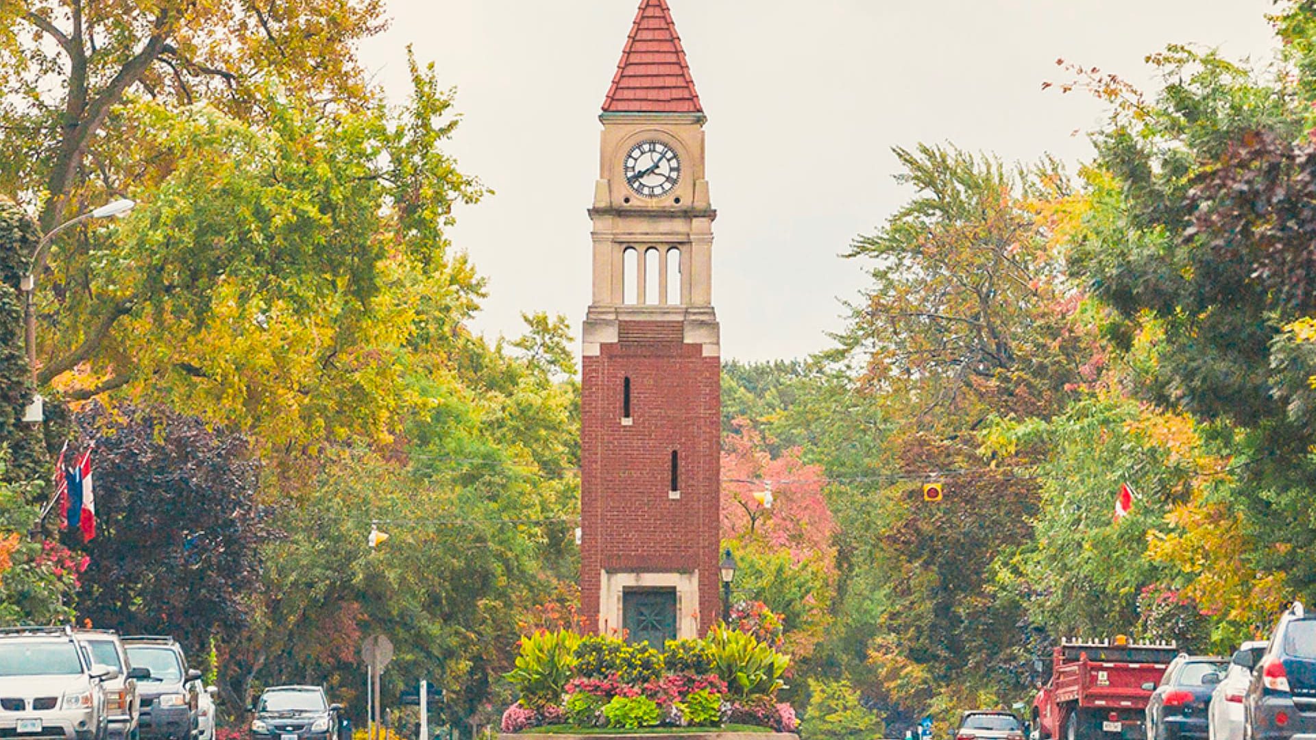 A brick clock tower with a red-tiled roof stands in the center of a tree-lined street, surrounded by cars and vibrant autumn foliage. There are colorful flowers planted at the tower’s base. 124 on Queen Hotel and Spa | Hotel & Spa Niagara-on-the-lake | 1 (855) 988-4552