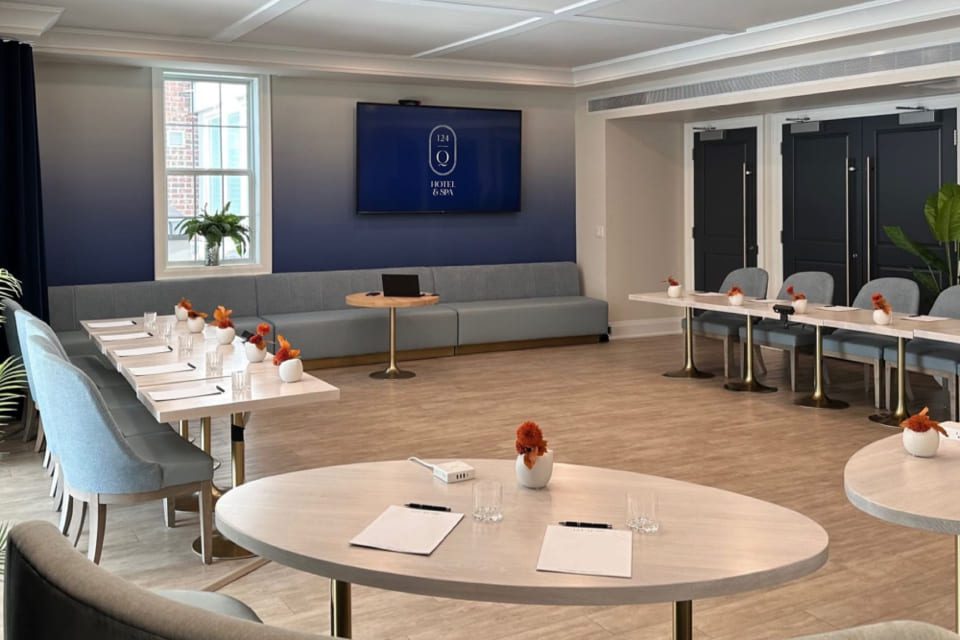 A modern meeting room with round and rectangular tables arranged with notepads, pens, and small vases of flowers, a gray sofa along the wall, large TV screen, and plenty of natural light from a window. 124 on Queen Hotel and Spa | Hotel & Spa Niagara-on-the-lake | 1 (855) 988-4552