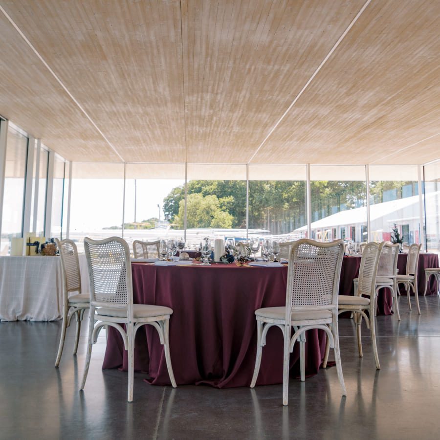 A round table with a burgundy tablecloth is set for a meal with glassware and white chairs in a bright, modern room with large windows overlooking a waterfront view. 124 on Queen Hotel and Spa | Hotel & Spa Niagara-on-the-lake | 1 (855) 988-4552