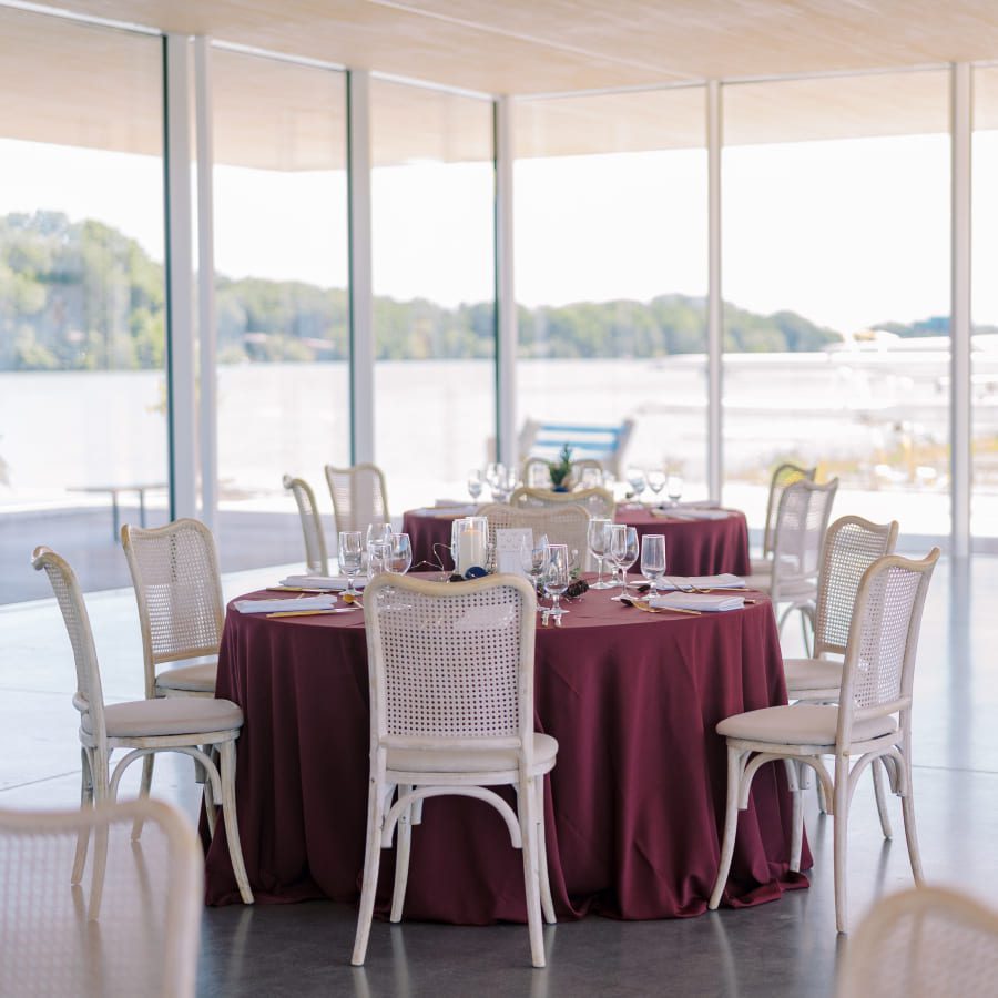 A round table with a burgundy tablecloth is set for a meal with glassware and white chairs in a bright, modern room with large windows overlooking a waterfront view. 124 on Queen Hotel and Spa | Hotel & Spa Niagara-on-the-lake | 1 (855) 988-4552