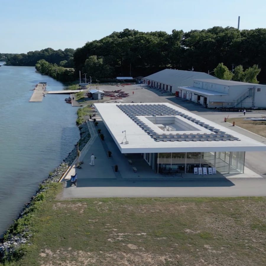A modern white building stands near the edge of a calm river, surrounded by trees and additional structures. A small boat is visible on the water under a clear blue sky. 124 on Queen Hotel and Spa | Hotel & Spa Niagara-on-the-lake | 1 (855) 988-4552