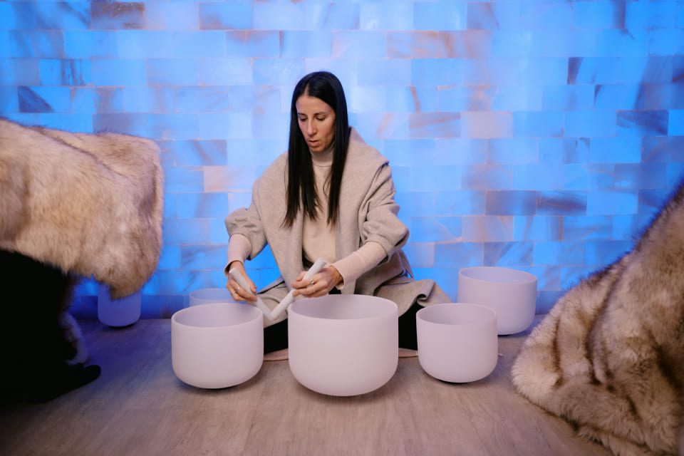 A woman sits cross-legged on the floor in front of several white crystal singing bowls, using mallets to play them. She is wrapped in a light-colored shawl, with blue-toned tiles and fur rugs around her. 124 on Queen Hotel and Spa | Hotel & Spa Niagara-on-the-lake | 1 (855) 988-4552 A woman sits cross-legged on the floor in front of several white crystal singing bowls, using mallets to play them. She is wrapped in a light-colored shawl, with blue-toned tiles and fur rugs around her. 124 on Queen Hotel and Spa | Hotel & Spa Niagara-on-the-lake | 1 (855) 988-4552
