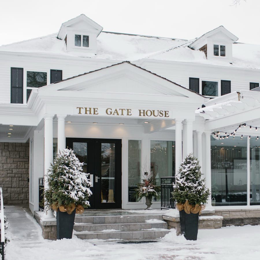 A white building with The Gate House sign above the entrance, decorated with potted plants and covered in snow. Steps lead to double doors, and string lights hang from a glass-enclosed area on the right. 124 on Queen Hotel and Spa | Hotel & Spa Niagara-on-the-lake | 1 (855) 988-4552