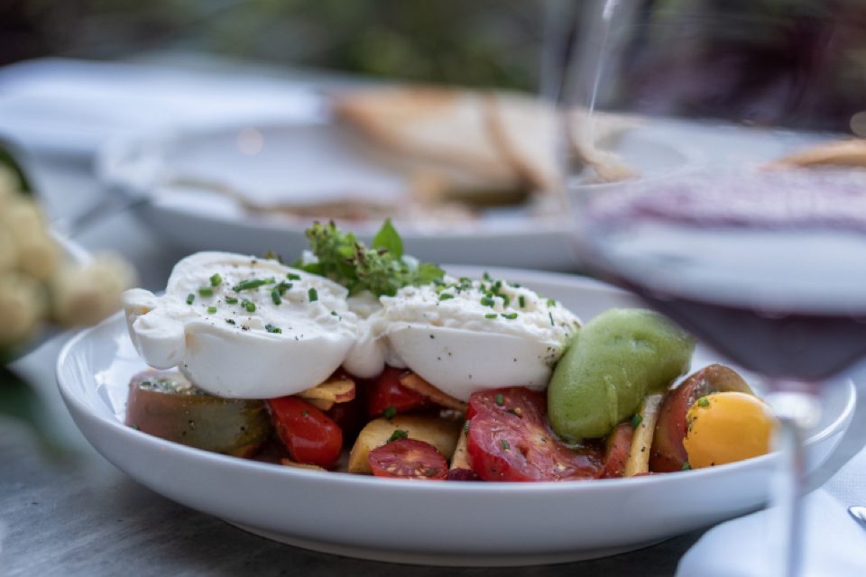 A close-up of a salad with fresh tomatoes, mozzarella cheese, green avocado, herbs, and chives on a white plate. A glass of red wine and some bread are blurred in the background. 124 on Queen Hotel and Spa | Hotel & Spa Niagara-on-the-lake | 1 (855) 988-4552