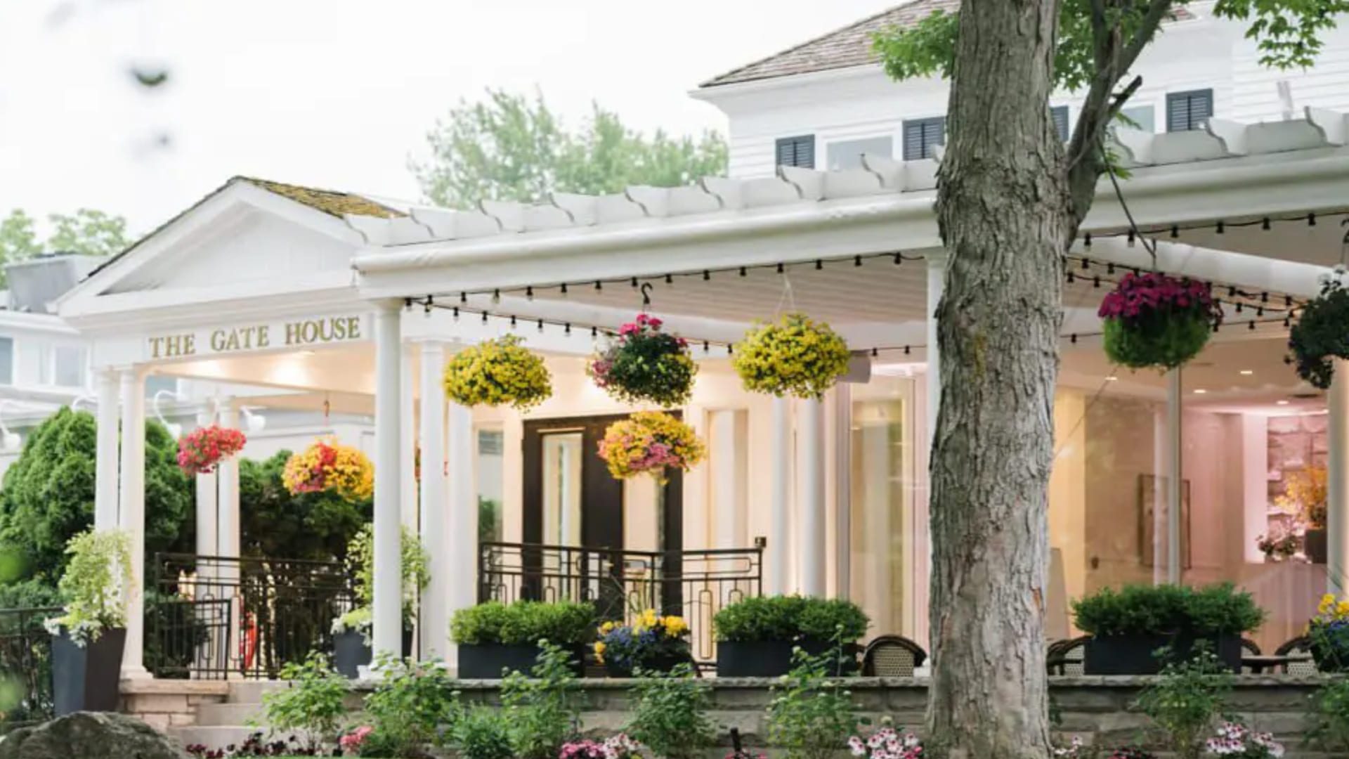 A charming white building labeled The Gate House features hanging flower baskets and string lights along a covered porch, surrounded by greenery and colorful gardens. 124 on Queen Hotel and Spa | Hotel & Spa Niagara-on-the-lake | 1 (855) 988-4552