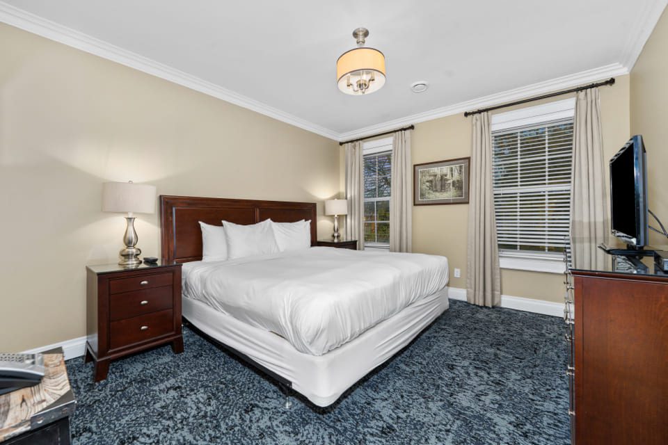 A neatly made bed with white linens is centered in a modern bedroom with beige walls, blue carpet, two nightstands with lamps, a TV on a dresser, large windows with blinds, and neutral decor. 124 on Queen Hotel and Spa | Hotel & Spa Niagara-on-the-lake | 1 (855) 988-4552