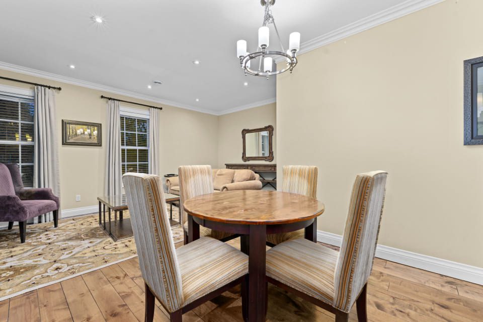 A dining area with a round wooden table and four striped upholstered chairs, adjacent to a living room with sofas, a rug, and large windows with curtains, all in a neutral color scheme. 124 on Queen Hotel and Spa | Hotel & Spa Niagara-on-the-lake | 1 (855) 988-4552